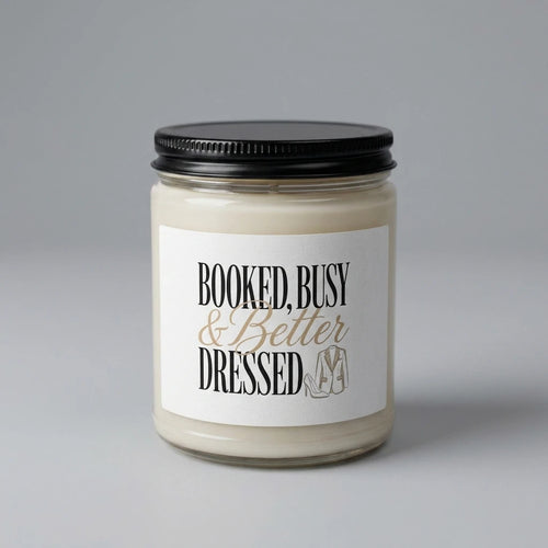 A soy candle in a glass jar with a label that reads Booked, Busy and Better Dressed.