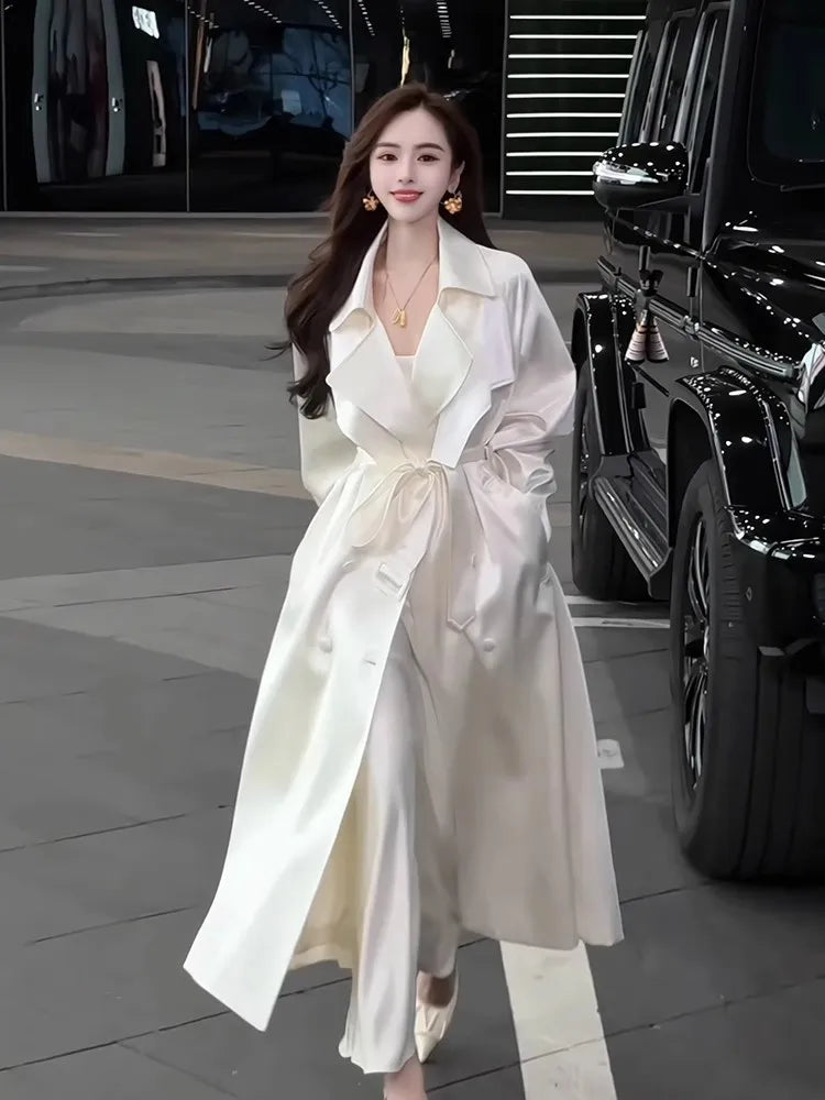 A woman wears a long, shiny white double-breasted trench coat with a sash belt.
