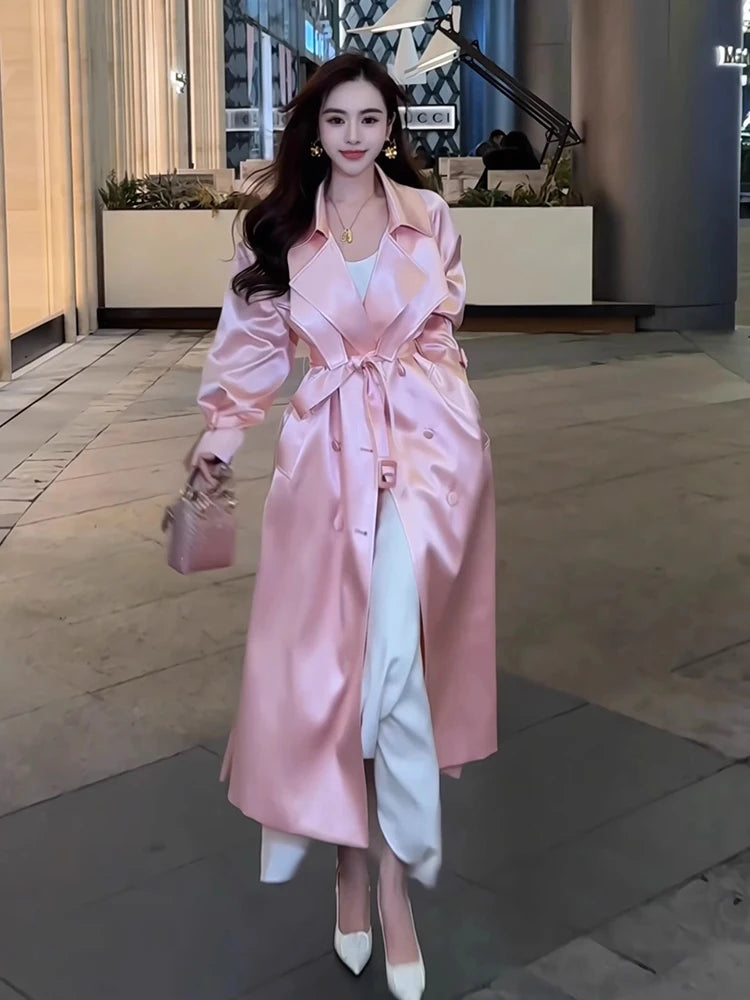 A woman wears a long, shiny pink double-breasted trench coat with a sash over white trousers.