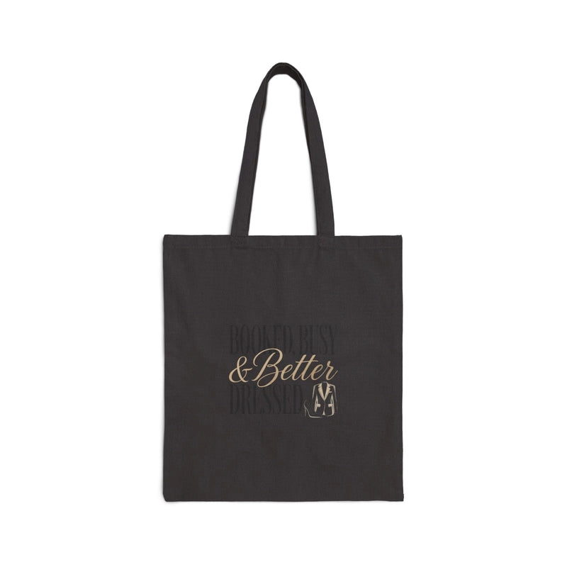 A black canvas tote bag featuring the text Booked, Busy and Better Dressed in gold lettering.